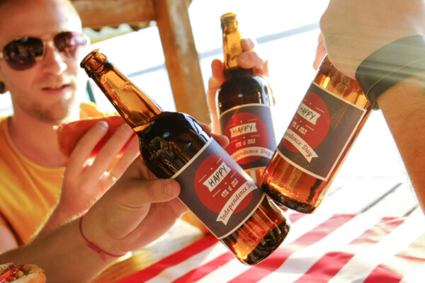 4th of July celebration party, young men hands holding beer bottle, holiday label design, Happy Independence day, hot dog, mustard, catchup. American flag, river recreation area.