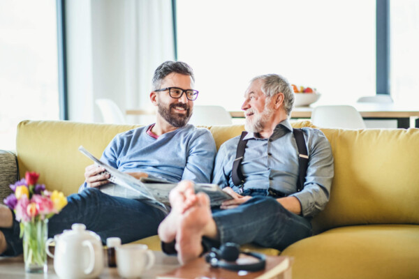 An adult son and senior father sitting on sofa indoors at home, talking.