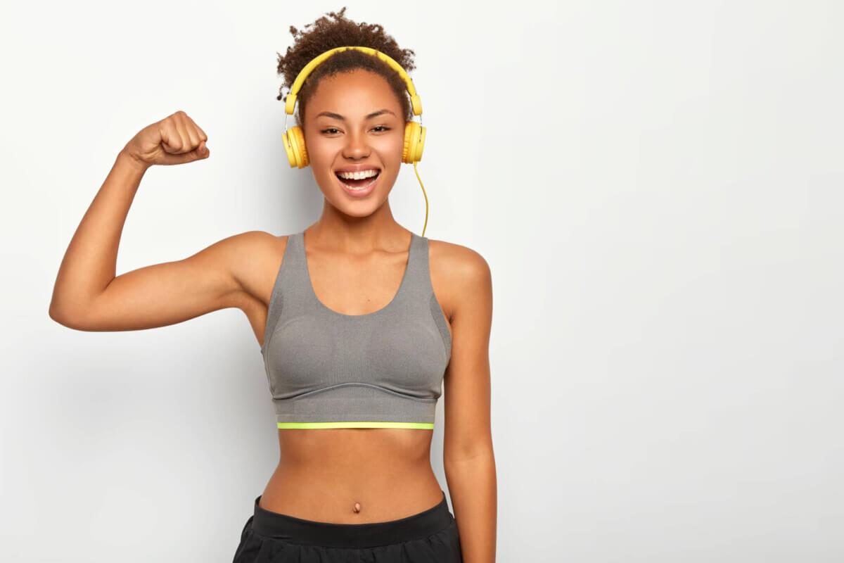 Horizontal view of dark skinned woman in good mood, raises arm with muscles, has strong body, dressed in gym outfit, listens audio via modern headphones, poses indoor. Fitness and music concept