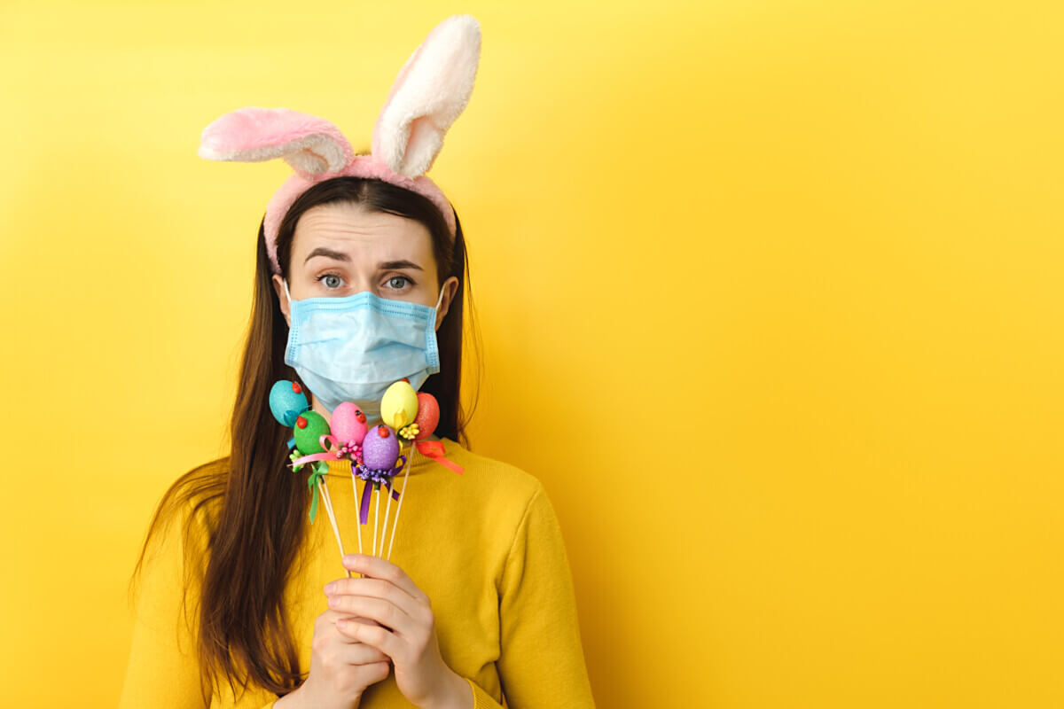 Discontent young woman with virus mask and looks unhappily, has spoiled day, poses with small Easter eggs, wears sweater, rabbit fluffy ears, poses. Concept holiday, coronavirus, virus protection