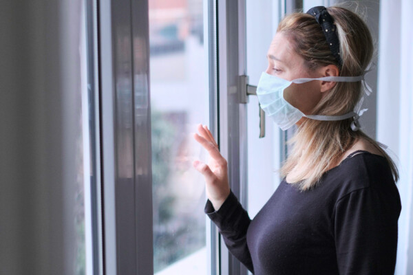 Woman wearing mask looking out window while under quarantine isolation for coronavirus / COVID-19 outbreak