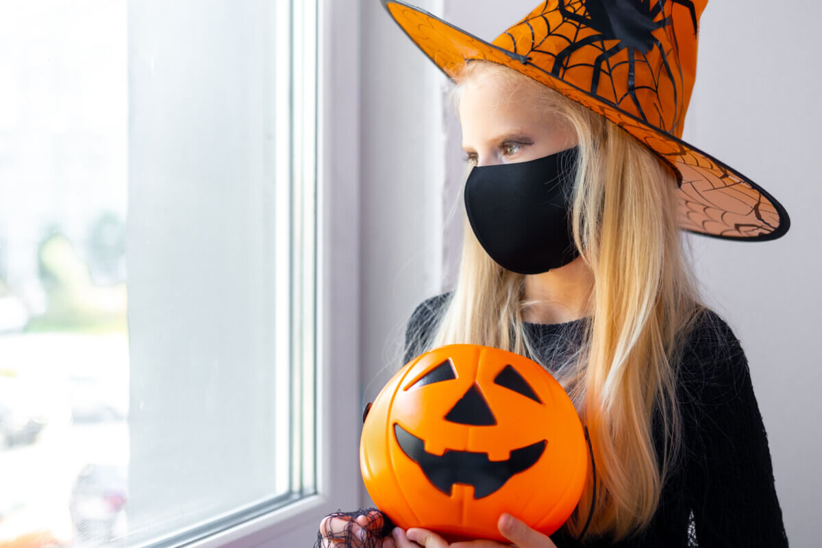 Sad thoughtful blonde girl in witch costume wearing mask looking in window at home with candy bucket. preparing for Halloween protecting from covid