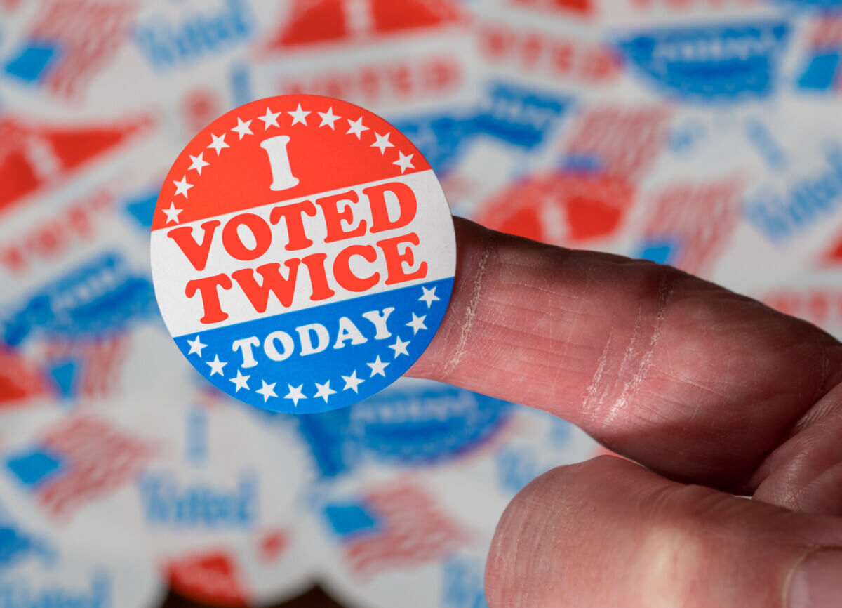 Finger with I Voted Twice sticker in front of many election voting badges to illustrate voter fraud
