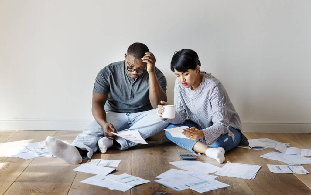 Couple sitting in pile of bills, as financial struggles, money worries, debt grow