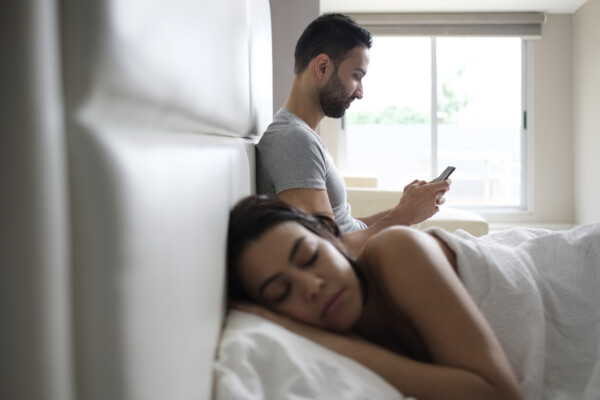 Man texting in bed on smartphone next to sleeping wife
