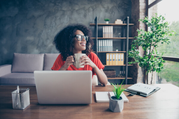 Happy woman drinking coffee at work desk, looking out office window smiling