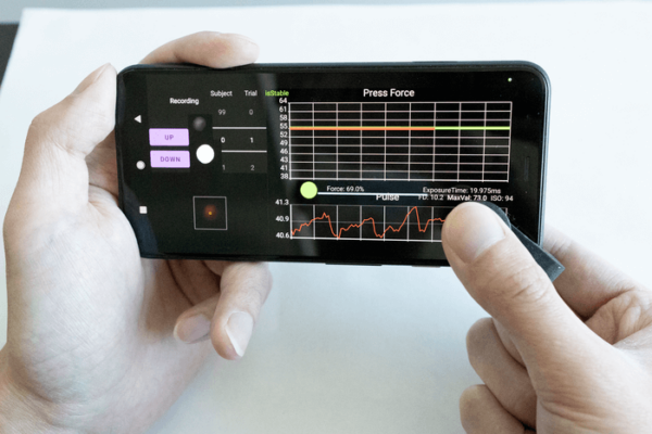 Two hands holding prototype of a blood pressure monitoring clip. Monitor screen show green, orange, and red, wavy horizontal lines