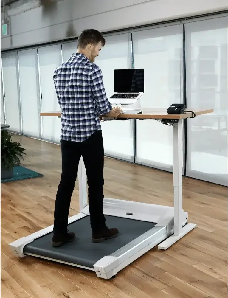 man in plaid shirt and jeans walks on treadmill with computer in front of him on desk