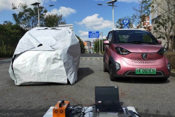 two cars in a parking lot, once covered by a thermal cloak, the other is a red SUV uncovered. In front the cars are devices that measure temperatures