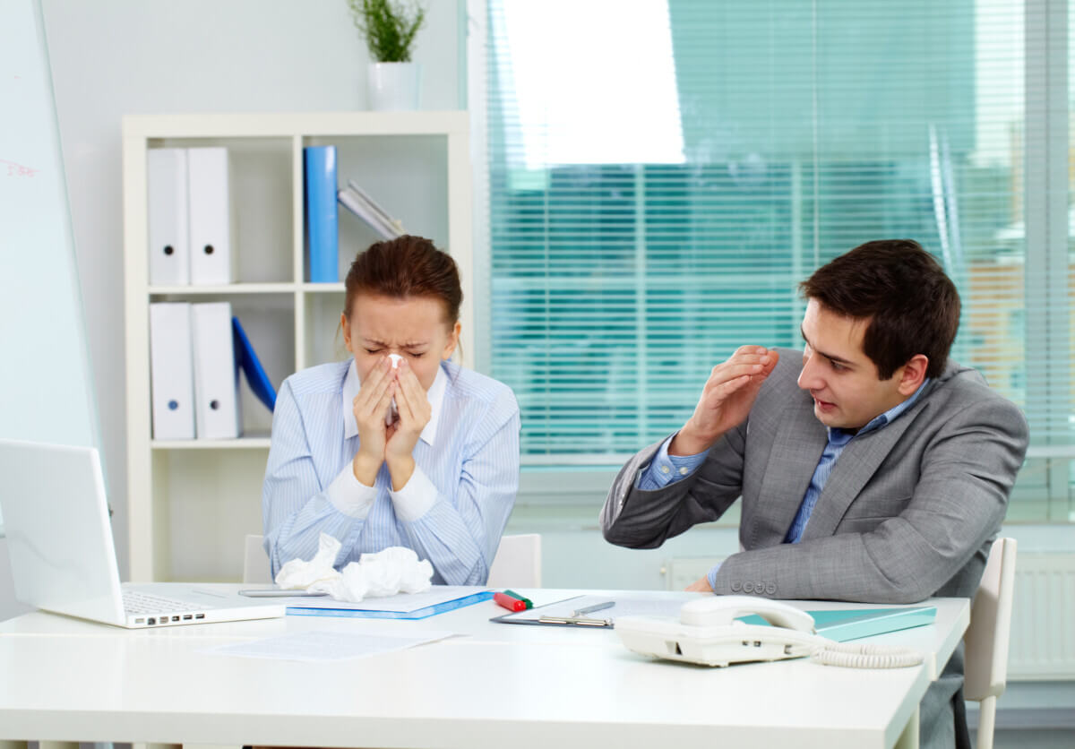 Man covers his face while his sick coworker blows her nose at the office.