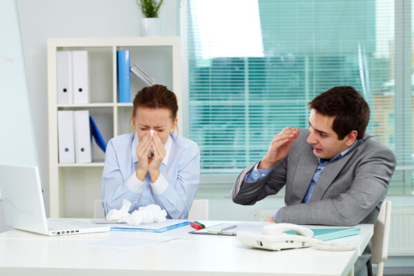 Man covers his face while his sick coworker blows her nose at the office.
