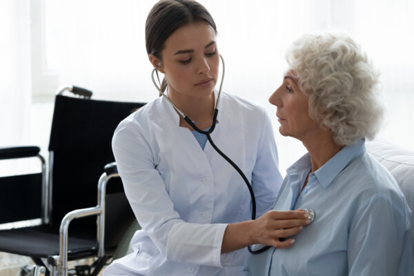 female physician caring for elderly woman on couch using stethoscope