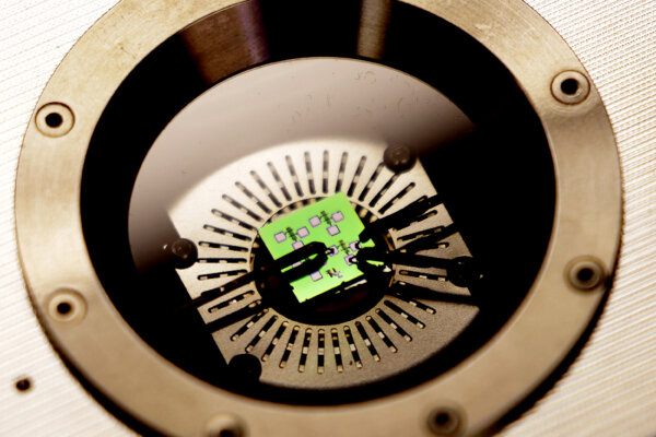 Black pins secure a green computer chip onto a metal platform inside a metal box. A circular window provides a view inside.