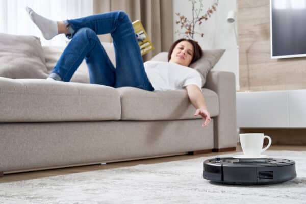 Robotic vacuum cleaner cleaning room while woman resting on sofa
