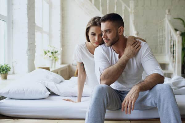 Depressed or sad man sitting in bed, being comforted by his wife or girlfriend