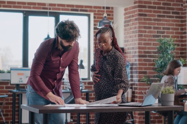 Coworkers, one white male, one Black pregnant woman in office looking at paperwork on a desk