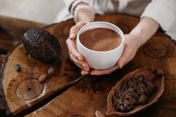 Hot handmade ceremonial cacao in white cup. Woman hands holding craft cocoa, top view on wooden table. Organic healthy chocolate drink prepared from beans, no sugar. Giving cup on ceremony, cozy caf