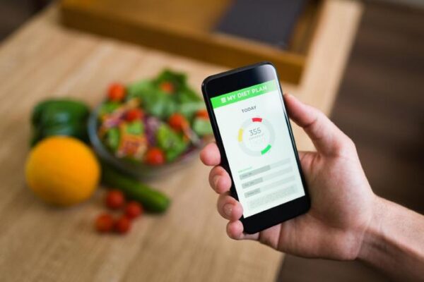 picture pf someone holding a phone with a food tracker open on the screen and fressh veggies on the table in the background