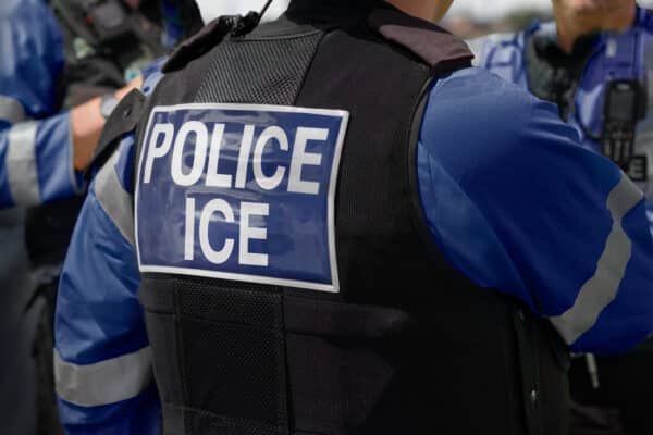 Ice Police Agent - officer of Immigration and Customs Enforcement. Close-up of POLICE ICE marking on the back of a stab proof vest worn by a trio of police officers standing around outdoors