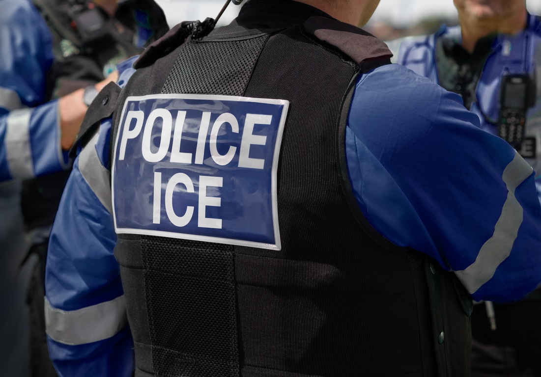 Ice Police Agent – officer of Immigration and Customs Enforcement. Close-up of POLICE ICE marking on the back of a stab proof vest worn by a trio of police officers at the scene of immigrant incident.