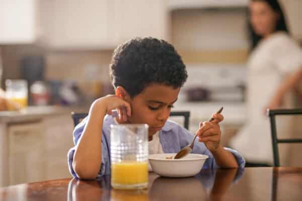boy eating breakfast with a spoon out of a bowl while sitting at a kitchen table. Table in the background standing at the stove looking at him