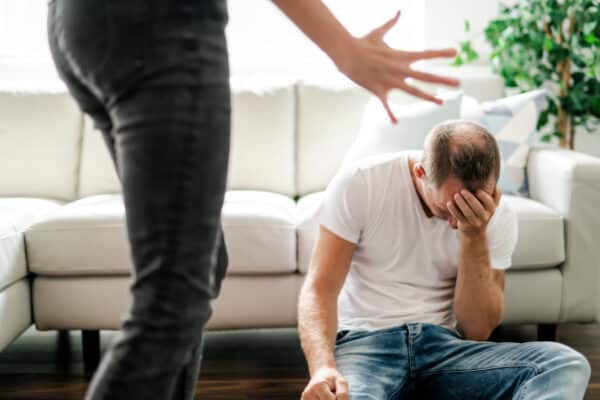 woman standing over a man aggressively as he sits on the floor with his head in his hand