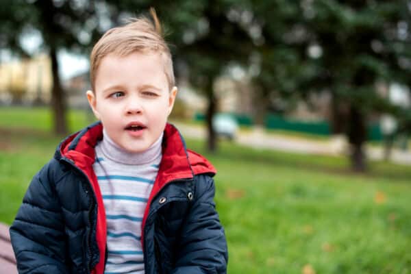 A boy with blond hair winks with one eye. He stands on a background of blurred green trees. The photo is blurred. High quality photo