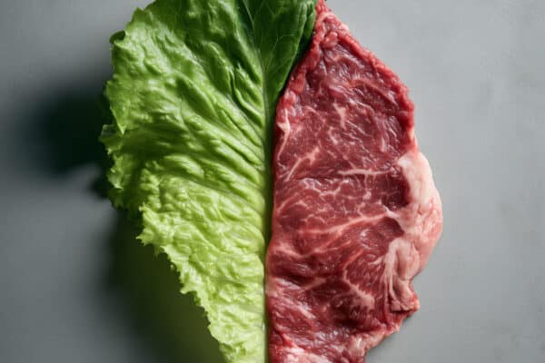 Creative split composition pairing a fresh green lettuce leaf with a raw beef steak on a neutral gray surface, representing nutrition, diet choices and food contrast.