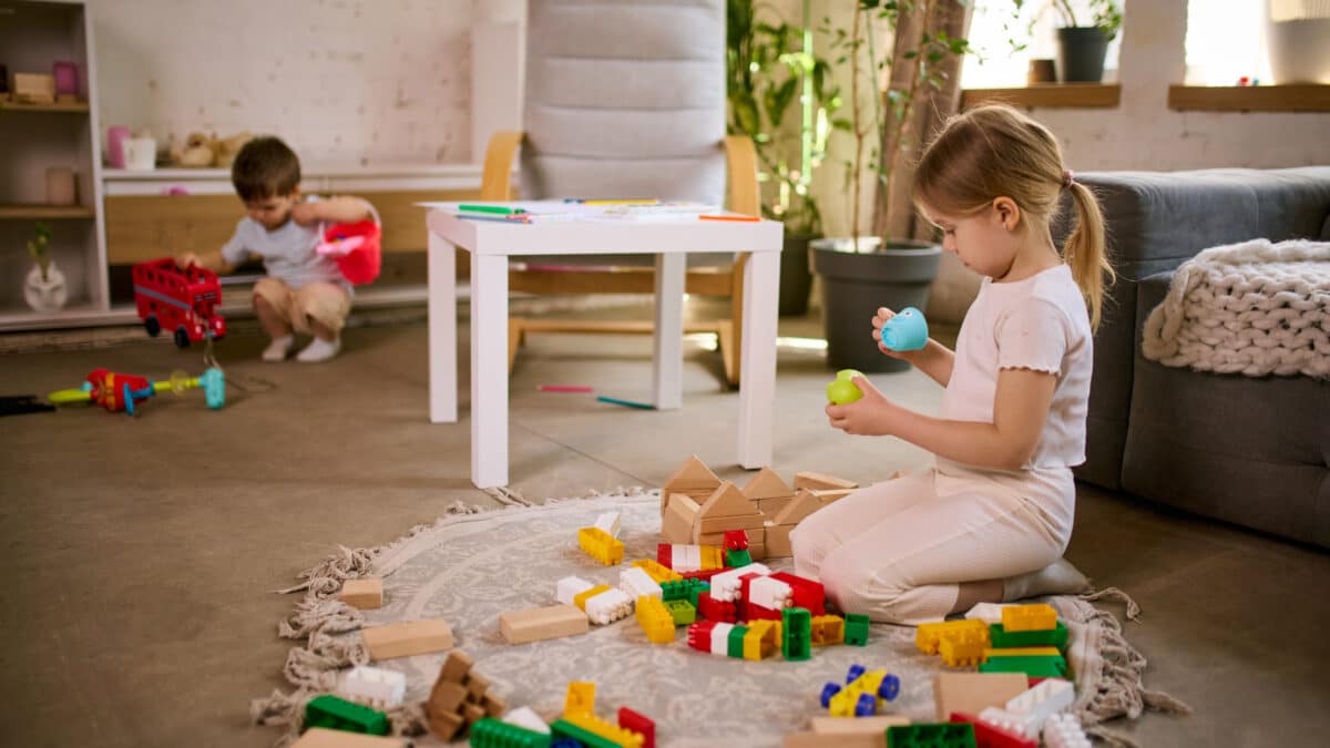 Little girl playing with toys while boy in background making mess in living room.