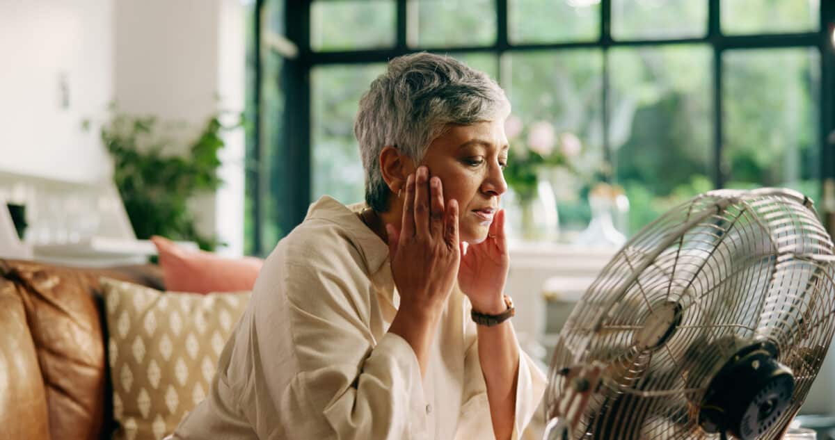 Senior woman, hot and fan at house for heatwave, air conditioning and menopause symptoms. Elderly person, sweat and electric appliance for cool breeze, warm weather and relief from hormonal condition