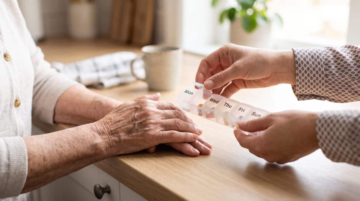 of caregiver hands giving pill organizer box to elderly woman, home healthcare assistance, senior citizen taking medication, weekly drug dispenser container, family support
