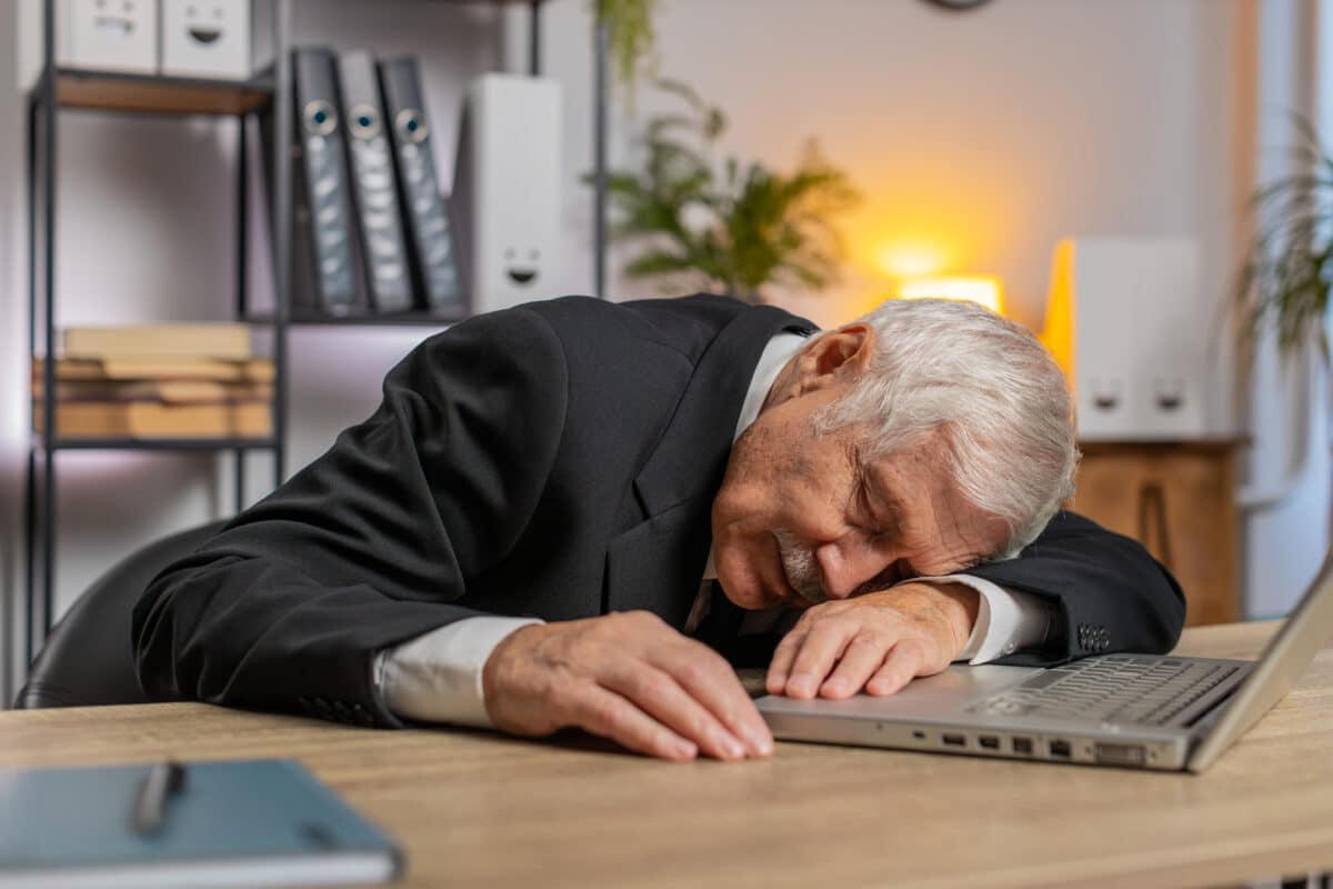 Bored sleepy senior businessman working on laptop leaning on hand falling asleep at office desk