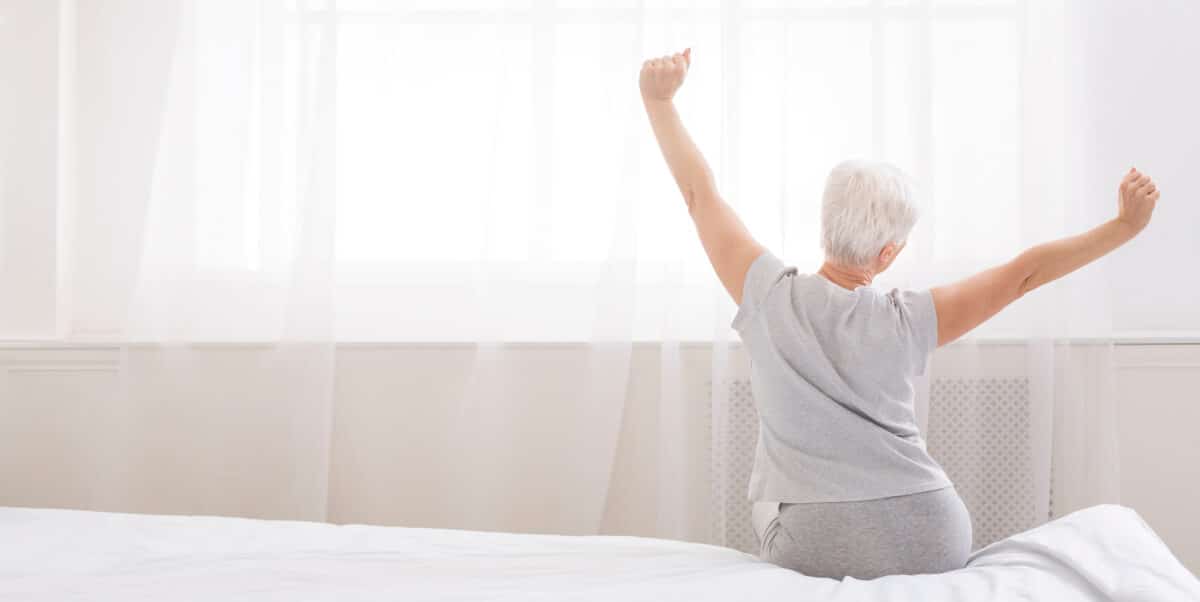 Well slept senior woman stretching with arms raised on bed