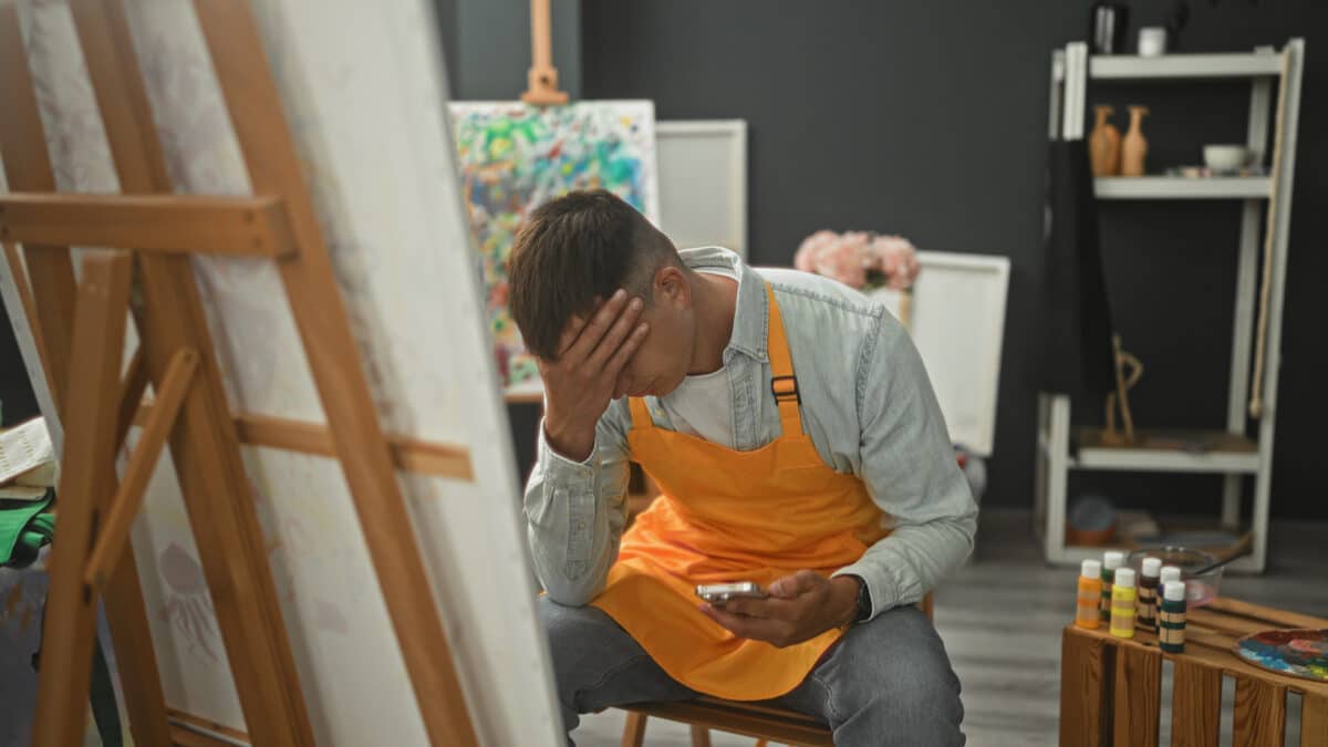 Man painter in studio sits on stool, holds smartphone with hand on forehead, wearing orange apron by easel and palette; creative frustration.