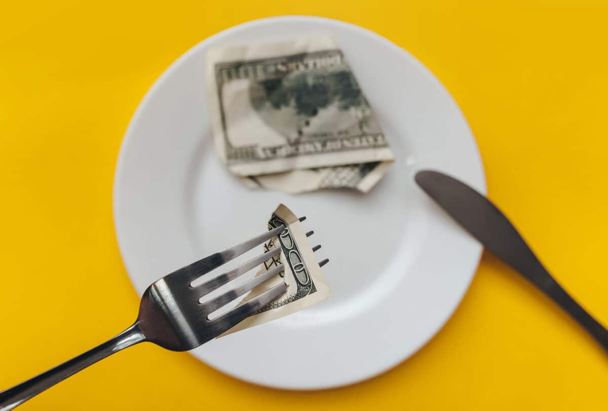 A slice dollar bill pierced with a fork. The knife and white plate on a yellow tablecloth. The concept of business lunch, banking, rentier life and financial well-being, income reduction.