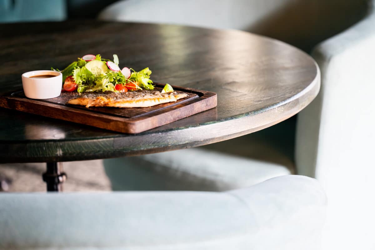 Pan-seared fish fillet served on a wooden board with a fresh green salad, cherry tomatoes, radishes, and a side of sauce on a dark round table in a restaurant setting